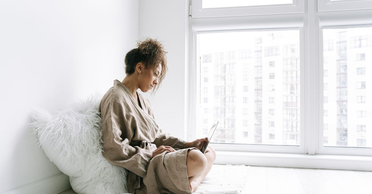 A woman sits on the floor beside a bright window in a calm, plain room.
