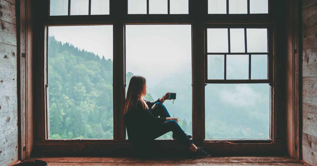 A woman sits alone in a large window frame looking out at the hills.